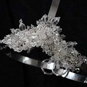 May include: A silver-toned bridal headpiece with clear crystal accents. The headpiece features a band that curves around the head, with a cluster of crystals at the top. The crystals are various shapes and sizes, adding sparkle and dimension.