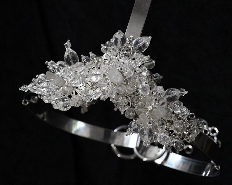 May include: A silver-toned bridal headpiece with clear crystal accents. The headpiece features a band that curves around the head, with a cluster of crystals at the top. The crystals are various shapes and sizes, adding sparkle and dimension.