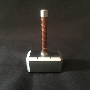 May include: A small silver-colored hammer with a brown leather wrapped handle. The hammer is a replica of Thor's hammer, Mjolnir.
