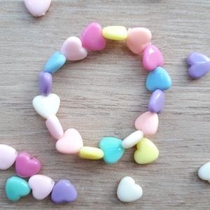 Peut inclure: Un bracelet composé de perles en forme de cœur aux couleurs pastel : rose, bleu, jaune et violet. Le bracelet est disposé en cercle, avec des perles en vrac éparpillées sur une surface en bois. Les perles mesurent environ 1 cm.