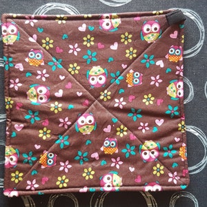 May include: Square brown quilted potholder with a repeating pattern of colorful owls, flowers, and hearts. The owls have large eyes and are in various colors. The potholder has a black loop for hanging.