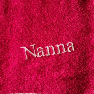 Puede incluir: Primer plano de una toalla rosa brillante con el nombre "Nanna" bordado en escritura blanca. La toalla tiene una superficie texturizada y un borde blanco en la parte inferior. La imagen destaca la textura suave y afelpada de la toalla.