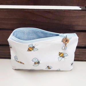 May include: A white zippered pouch with a blue lining and a small gold charm. The pouch is decorated with a repeating pattern of bees.