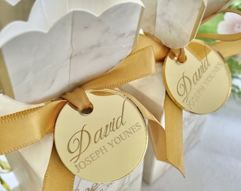 May include: Two white favor boxes with gold ribbon and gold round tags. The tags have the name "David Joseph Younes" engraved on them.