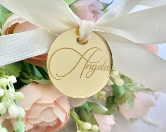 May include: Gold infinity symbol with the name "Angela" engraved on a round, gold mirror tag attached to a white satin ribbon.