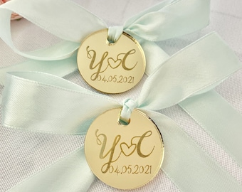 May include: Two gold round acrylic wedding favor tags with a light blue ribbon. The tags are engraved with the letters Y and C with a heart between them and the date 04.05.2021.