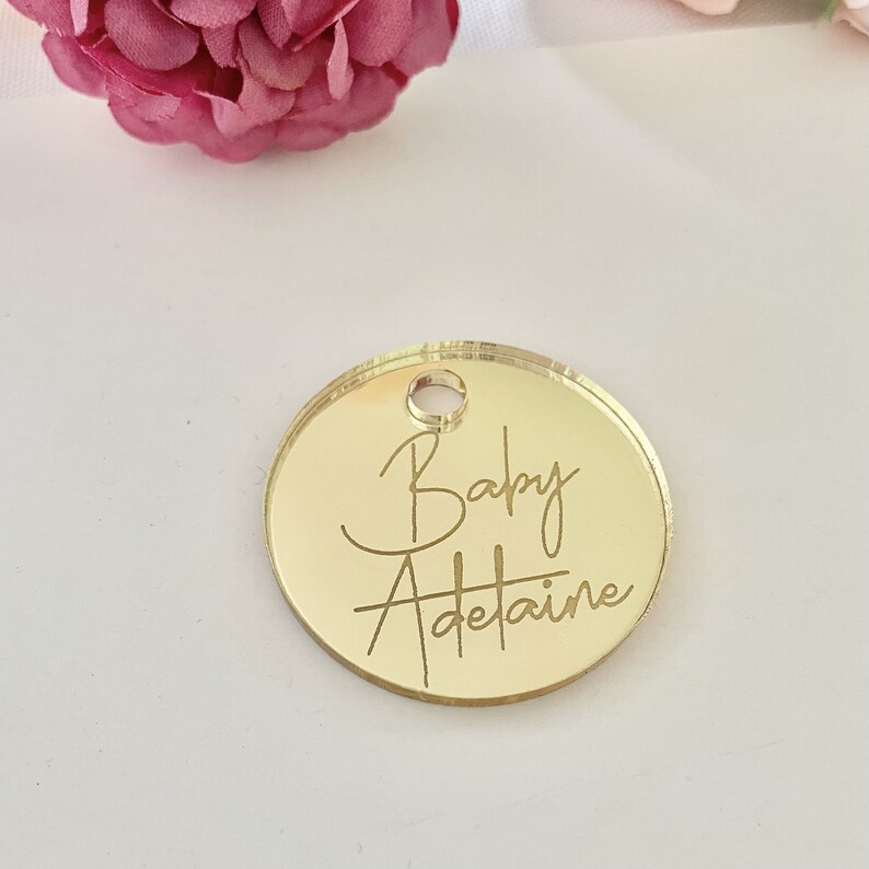 May include: Gold round mirror tag with the text "Baby Adetaine" engraved in a cursive font.