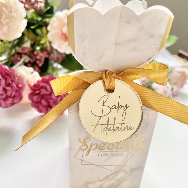 May include: Gold favor box with a white marble pattern and a gold ribbon. A gold circle tag with the text "Baby Adetaine" is attached to the ribbon. The box has the text "Specially for you!" printed on the front.