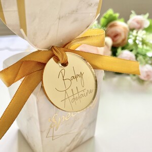 May include: A gold round favor tag with the text "Baby Adelaide" written in cursive script. The tag is tied to a white and gold gift box with a yellow ribbon.