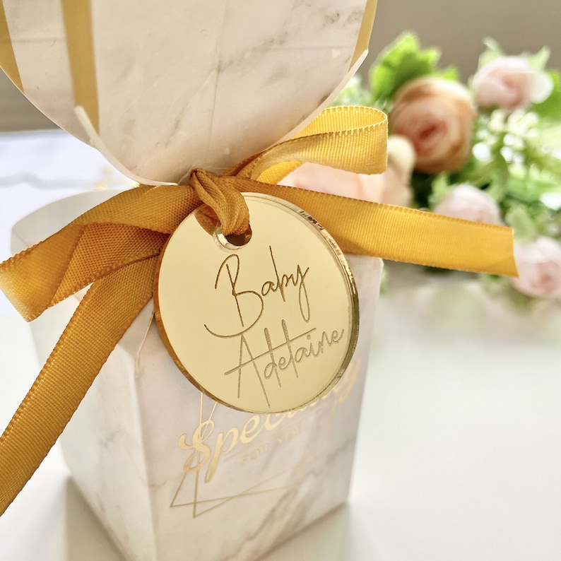 May include: A gold round favor tag with the text "Baby Adelaide" written in cursive script. The tag is tied to a white and gold gift box with a yellow ribbon.