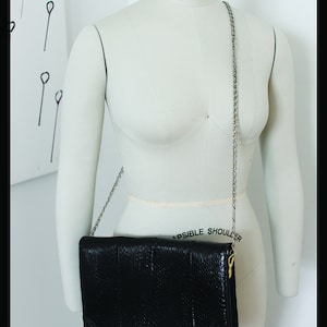 Könnte beinhalten: Eine schwarze Leder-Clutch mit einem silbernen Kettenriemen. Die Tasche hat eine strukturierte Oberfläche und einen goldenen Verschluss.