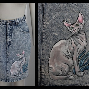 Può includere: Una gonna di jeans con un designo dipinto a mano di un gatto senza pelo. Il gatto è seduto con la coda arrotolata intorno alle zampe. La gonna è di colore blu chiaro con un aspetto sbiadito.