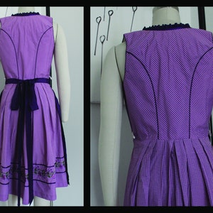May include: A sleeveless purple dirndl dress with a checkered pattern. The dress features a dark purple trim, a matching sash, and floral embroidery at the hem. The dress is displayed on a mannequin.