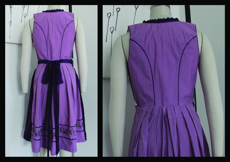 May include: A sleeveless purple dirndl dress with a checkered pattern. The dress features a dark purple trim, a matching sash, and floral embroidery at the hem. The dress is displayed on a mannequin.