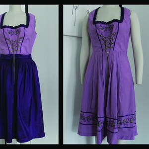 May include: A sleeveless purple dirndl dress with a checkered bodice and a solid purple skirt. The bodice features a square neckline, decorative lacing, and floral embroidery. The dress is displayed on a mannequin.