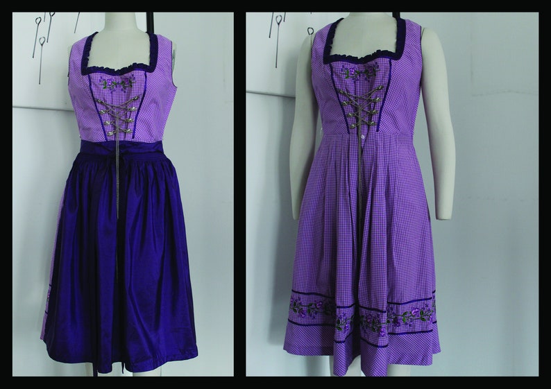 May include: A sleeveless purple dirndl dress with a checkered bodice and a solid purple skirt. The bodice features a square neckline, decorative lacing, and floral embroidery. The dress is displayed on a mannequin.