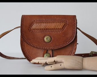 May include: A brown leather satchel bag with a flap closure and a decorative stitched design. The bag has a long adjustable strap and two metal button closures.