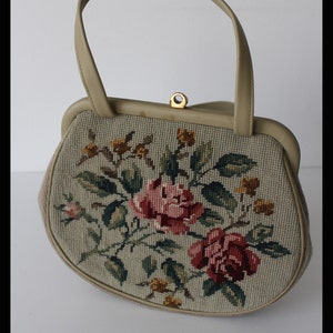 May include: A beige handbag with a floral design. The bag is made of a tapestry fabric with a floral pattern in shades of pink, green, and yellow. The bag has a top handle and a clasp closure.