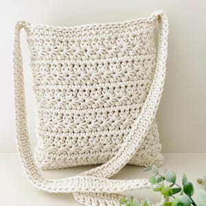 May include: A crocheted cream-colored shoulder bag with a long strap. The bag is made with a simple stitch pattern and has a slightly textured surface.