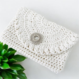 May include: A white crocheted clutch bag with a silver button. The bag is made with a simple stitch pattern and has a rounded flap.