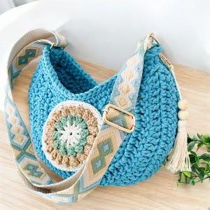 May include: A turquoise crochet crossbody bag with a patterned strap and a decorative circular patch. The strap is beige with blue and tan geometric designs. The bag has a tassel and wooden beads.