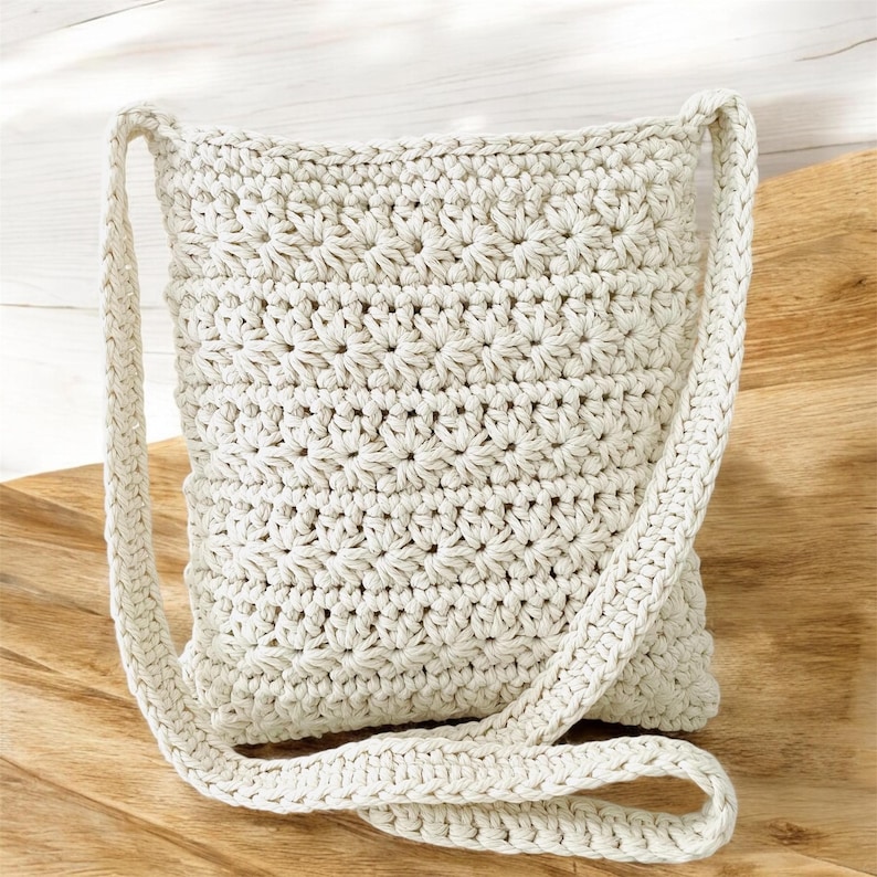 May include: A cream-colored crocheted crossbody bag with a long strap. The bag is made with a simple stitch pattern and has a slightly textured surface.