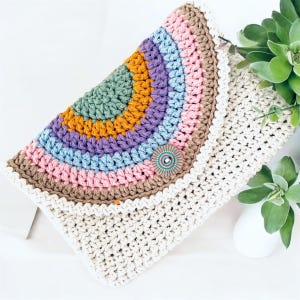 May include: A handmade crochet clutch in a rainbow design. The top flap features a semi-circle of colors including green, orange, purple, blue, and pink. The clutch is secured with a decorative button. The body of the clutch is off-white.