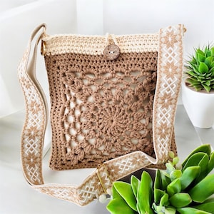 May include: A handmade crochet shoulder bag in beige and brown. The bag features a detailed floral pattern on the front and a decorative strap with a geometric design. A wooden button secures the flap. The bag is displayed with a small potted plant.