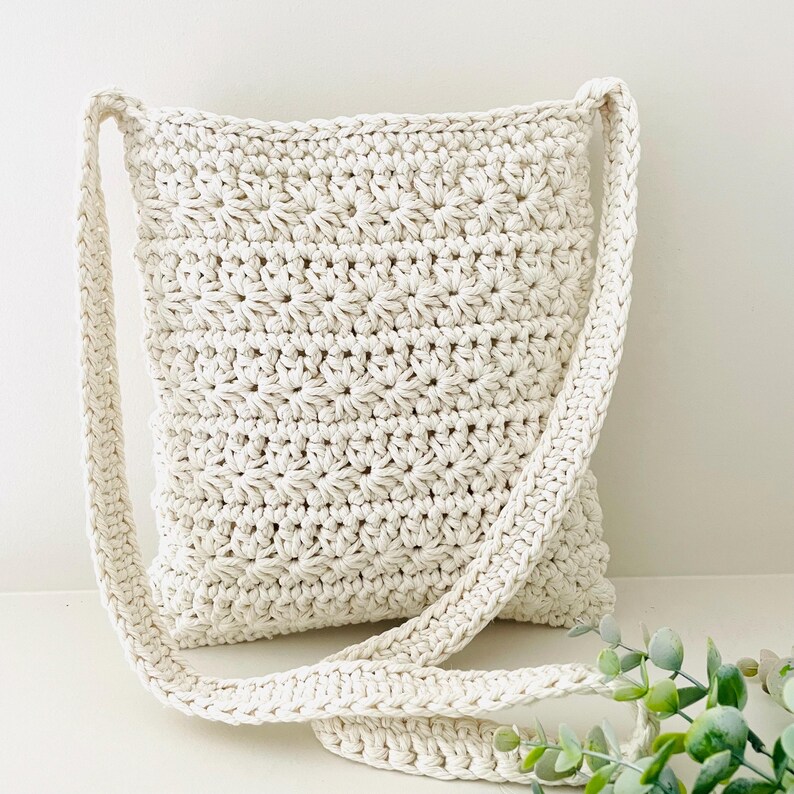 May include: A crocheted cream-colored crossbody bag with a long strap. The bag is made with a simple stitch pattern and has a relaxed, casual look.