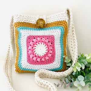 May include: A crocheted crossbody bag with a colorful geometric pattern in shades of pink, white, blue, and yellow. The bag has a long strap and a wooden button closure.