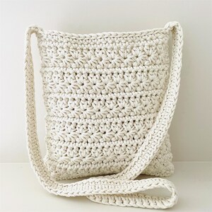 May include: A white crocheted crossbody bag with a long strap. The bag is made with a simple stitch pattern and has a relaxed, casual look.