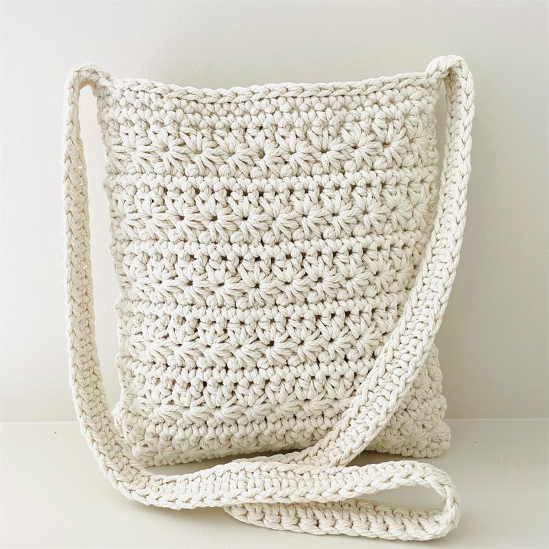 May include: A white crocheted crossbody bag with a long strap. The bag is made with a simple stitch pattern and has a relaxed, casual look.