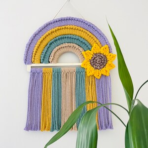 May include: A colorful macrame rainbow wall hanging with a crocheted sunflower in the center. The rainbow is made of purple, yellow, green, and beige yarn. The sunflower is yellow with a brown center.