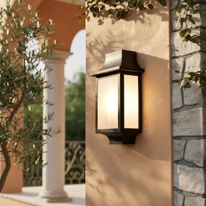 Puede incluir: Un aplique de pared rectangular para exteriores con marco de metal negro mate y paneles de vidrio esmerilado. La lámpara presenta un diseño escalonado en la parte superior e inferior, emitiendo un brillo cálido y suave, montada en una pared clara.