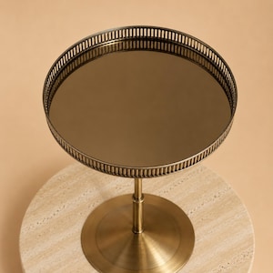 Puede incluir: Una bandeja redonda con pedestal, superficie de espejo oscuro y borde de metal color bronce con una barandilla de listones verticales. La bandeja se apoya en una columna central delgada y una base circular ancha con acabado metálico.