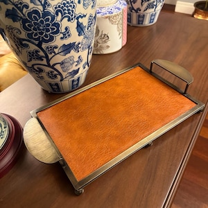 May include: A rectangular serving tray featuring a textured, burnt orange faux leather base set within a metallic, antique brass-toned frame. The tray includes two curved side handles and is supported by four small, rounded feet at the corners.