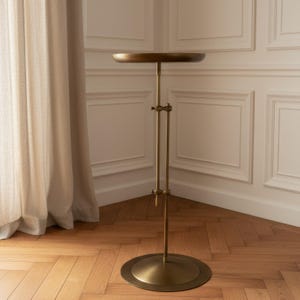 May include: A tall, adjustable metal pedestal stand with a circular top and a matching circular base. The stand features a central vertical pole with a sliding mechanism and a locking screw, finished in a muted, brushed antique brass tone.