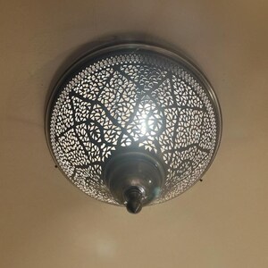 May include: A decorative ceiling light fixture with a detailed, openwork metal shade. The shade has a pattern of small, geometric shapes that allow light to filter through. The fixture is mounted on a neutral-colored ceiling, creating a warm ambiance.