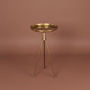 May include: A gold-toned metal side table featuring a circular tray-style top with a raised edge. The table is supported by three slender, straight legs that meet at a central point beneath the tabletop, creating a tripod base structure.