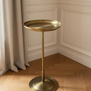 May include: A brass-toned metal pedestal table featuring a circular tray-style top with a raised edge, a slender central support column, and a wide, flat circular base. The surface has a brushed metallic finish with a warm golden hue.
