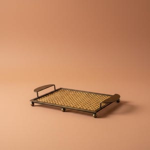 Op de afbeelding: Een rechthoekig dienblad met een basis van gevlochten riet, omlijst door een donkerbronzen metalen rand. Het dienblad is voorzien van twee gebogen metalen handgrepen aan de uiteinden en rust op kleine, bolvormige metalen voetjes aan de onderzijde.