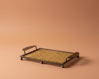 Puede incluir: Una bandeja rectangular con una base de rejilla tejida enmarcada por un borde de metal color bronce oscuro. La bandeja incluye dos asas metálicas curvas en los extremos y se apoya sobre pequeños pies esféricos de metal en cada esquina.