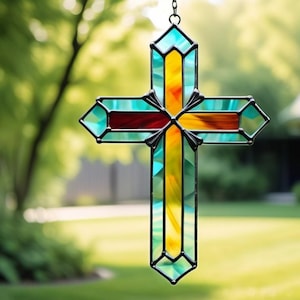 Puede incluir: Una cruz de vidrieras con un marco de metal negro cuelga sobre un fondo verde borroso. La cruz presenta colores vibrantes, incluyendo turquesa, amarillo y rojo, creando un efecto visual llamativo. La cruz está suspendida por una cadena.
