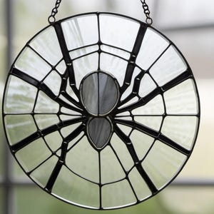 May include: A circular stained glass suncatcher featuring a black spider centred on a clear, textured web design. The spider has a two-part body made of opaque white glass, and the entire piece is framed in a thin black metal border with a chain.