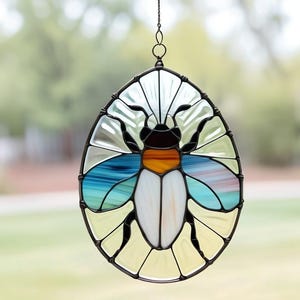 May include: Stained glass suncatcher in an oval shape, featuring a stylized beetle design. The insect has a white body, blue and orange accents, and black details. The surrounding glass is a mix of white, yellow, and clear panels, all framed in black.