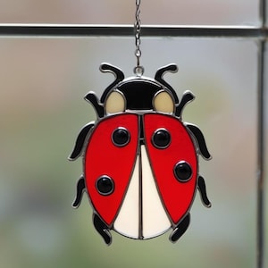 May include: A stained glass ladybug suncatcher with a silver-colored metal outline. The ladybug features a red body with black spots, black wings, and a black head. The ladybug is hanging from a silver chain.