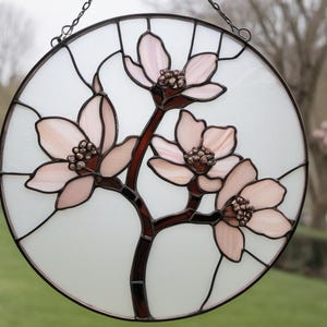 May include: Round stained glass window art featuring a floral design. The artwork showcases pink blossoms with brown centers and stems against a white background. The glass is framed with black lines and a metal chain for hanging.