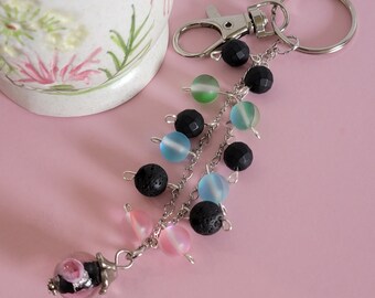 May include: A silver key chain with a charm featuring a pink and white glass bead. The chain is adorned with black lava rock beads and iridescent blue, green, and pink glass beads.