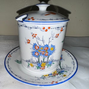 Puede incluir: Tarro de cerámica blanca con tapa y platillo a juego. El tarro y el platillo están decorados con diseños florales azules, naranjas y amarillos. El tarro tiene un pequeño asa para una cuchara. El borde del tarro y del platillo es azul.