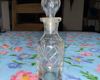 May include: A clear glass bottle with a diamond-shaped pattern and a clear glass stopper. The bottle is sitting on a blue and white floral tablecloth.